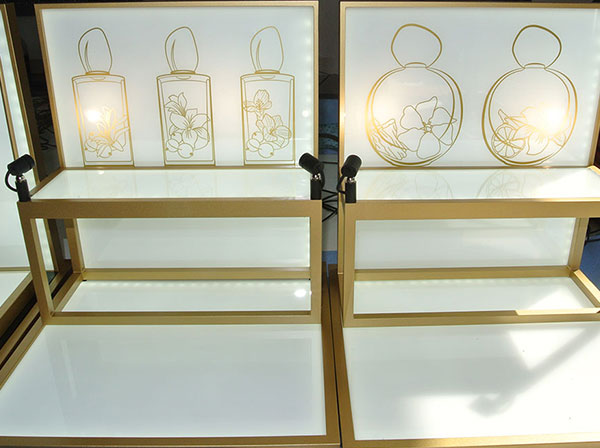 Produktdisplays aus Acrylglas in weiß mit goldenem Rand und gemalten Produktbildern im Hintergrund in Treppenform mit direkter und indirekter LED-Beleuchtung