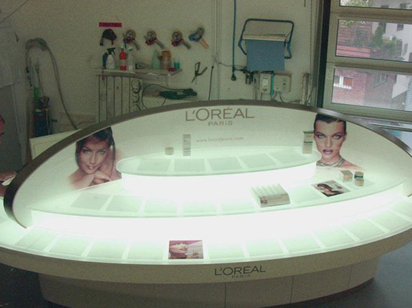 Ein ovaler Tisch aus weißem Acrylglas mit LED-Beleuchtung und mehrstufigem Produktdisplay für Kosmetikartikel für L`Oréal