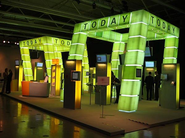 Messestand mit schrägen grünen und gelben Acrylglassäulen mit Beleuchtung, Beschriftung „today“ und montierten Bildschirmen